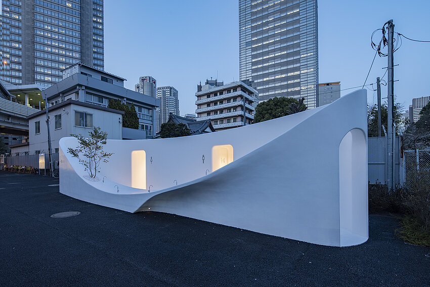 A white toilet house. The front wall is curved. The lowest point of the wall is only near the floor. Behind it are the entrances of the toilets. 