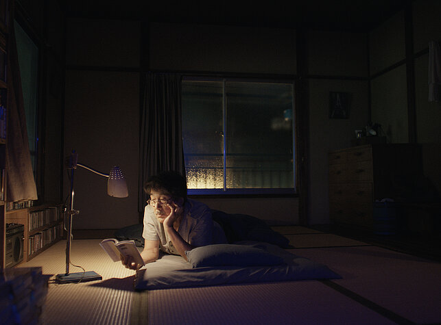 Hirayama is lying on a futon reading a book.