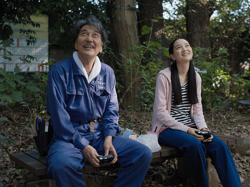 Hirayama and his niece Niko sit on a bench and look up smiling. 01_PerfectDays_Koji_Yakusho_Arisa_Nakano__c_2023_MASTER_MIND_Ltd.jpg